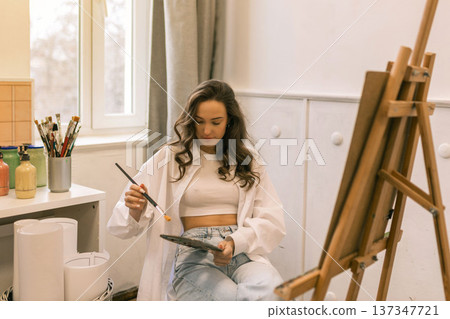 Young female artist standing near easel and holding pencil while working on painting in art studio, creative process and inspiration Young female artist standing near easel and holding pencil while working on painting in art studio, creative process and inspiration 137347721