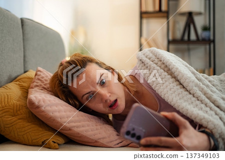 Woman feeling shocked reading bad news on phone in bed Woman feeling shocked reading bad news on phone in bed 137349103