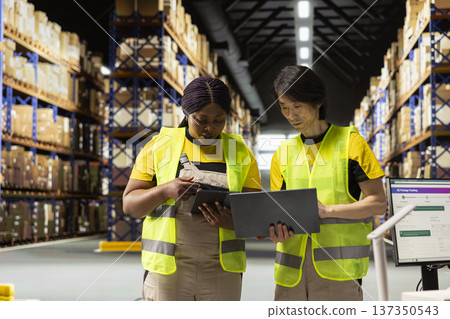 Multiethnic staff processing e-commerce orders and reviewing invoices, organizing cargo goods inside industrial warehouse space filled with storage racks. Smooth delivery and quality control. Multiethnic staff processing e-commerce orders and reviewing invoices, organizing cargo goods inside industrial warehouse space filled with storage racks. Smooth delivery and quality control. 137350543