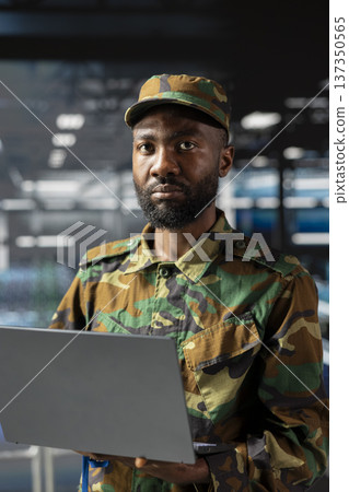 African american officer in server farm reviewing satellite footage on laptop, standing on industrial platform. Army personnel in high tech space doing reconnaissance operations via drone control. 137350565