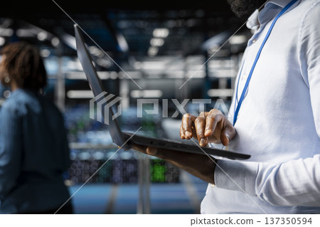 Close up of data center software engineer doing maintenance work on laptop, ensuring optimal performance. Male worker reviewing diagnostics and debugging server hub high tech equipment. Close up of data center software engineer doing maintenance work on laptop, ensuring optimal performance. Male worker reviewing diagnostics and debugging server hub high tech equipment. 137350594