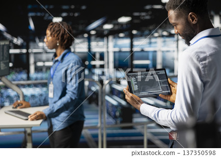 Server room IT programmer visualizing large language model infrastructure using tablet on elevated industrial platform. Data center system administrators collaborating on tasks. 137350598