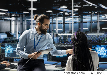 Data center team leader overseeing woman during internship running AI forecasting simulations. Server hub mentor guiding young female engineer integrating machine learning APIs into software 137350599