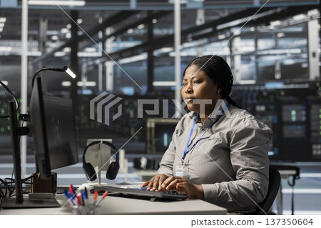 Data center software developer uses PC to update servers tech, configuring gear using program. Woman working in server farm reviewing hardware analytics, ensuring reliable network operations 137350604