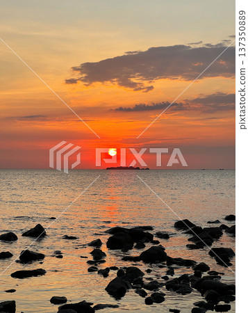 Vibrant sunset over calm water, silhouette of small island in the distance and dark rocks in the foreground. The sun is sinking towards horizon, turning the sky into warm shades of orange and red. 137350889