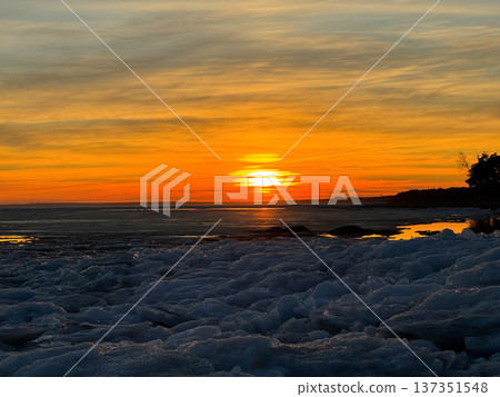 Serene winter sunset over frozen body of water, likely a lake or sea, with large chunks of blue and white ice covering foreground. Silhouetted trees line the distant shore, creating natural contrast. Serene winter sunset over frozen body of water, likely a lake or sea, with large chunks of blue and white ice covering foreground. Silhouetted trees line the distant shore, creating natural contrast. 137351548