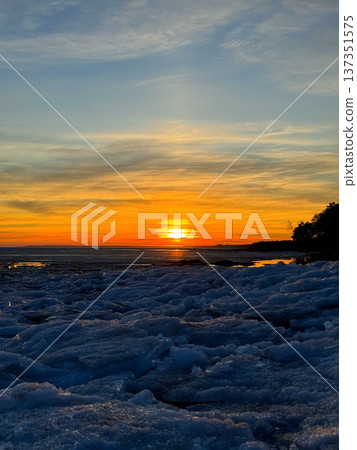 Fiery winter sunset over frozen body of water, likely a lake or sea, with large chunks of blue and white ice covering foreground. Silhouetted trees line the distant shore, creating natural contrast. 137351575