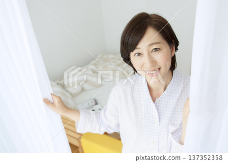 A middle-aged woman in pajamas opening the curtains A middle-aged woman in pajamas opening the curtains 137352358