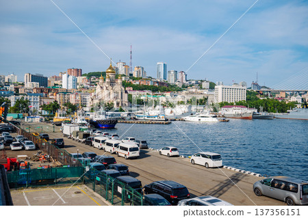 Vladivostok harbor bustling with parked vehicles, maritime vessels, urban landscape featuring golden church domes gleaming under bright sunlight Vladivostok harbor bustling with parked vehicles, maritime vessels, urban landscape featuring golden church domes gleaming under bright sunlight 137356151