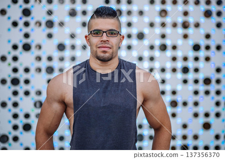 Muscular sportsman standing in front of a modern design wall, wearing a tank top and glasses Muscular sportsman standing in front of a modern design wall, wearing a tank top and glasses 137356370