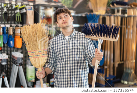 Guy in hardware store examines broom and besom. Guy in hardware store examines broom and besom. 137356574