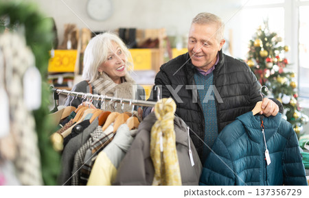 Elderly couple standing in Christmas shop and choosing jackets Elderly couple standing in Christmas shop and choosing jackets 137356729