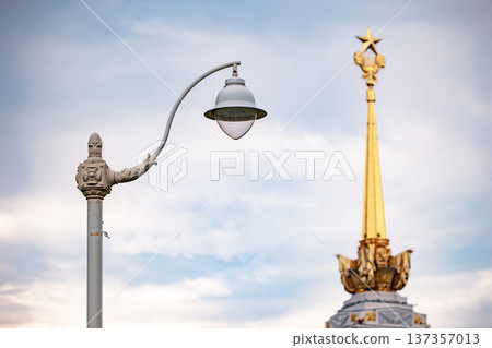 VDNH golden spire with red star and historic street lamp against cloudy sky VDNH golden spire with red star and historic street lamp against cloudy sky 137357013