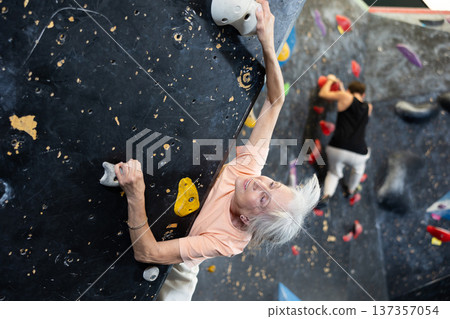 Sporty elderly female exercising in boulder climbing hall without rope, reaching new results, enjoying new challenges in amusement park Sporty elderly female exercising in boulder climbing hall without rope, reaching new results, enjoying new challenges in amusement park 137357054