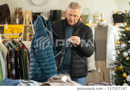 mature man chooses a down jacket against the background of a Christmas tree mature man chooses a down jacket against the background of a Christmas tree 137357197