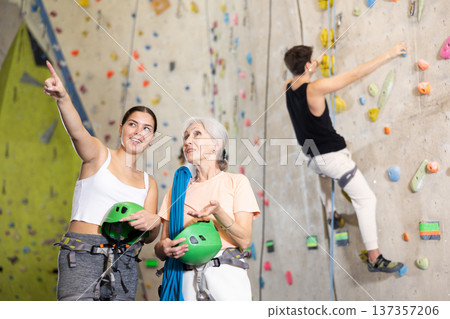 Young female trainer giving instructions to elderly woman about climbing artificial training rock wall in indoor bouldering gym Young female trainer giving instructions to elderly woman about climbing artificial training rock wall in indoor bouldering gym 137357206