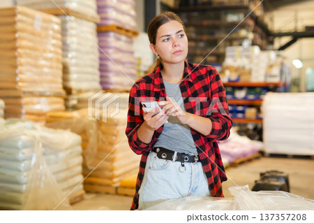 Female manager keeps records of bulk construction materials in the warehouse, writing down data on smartphone Female manager keeps records of bulk construction materials in the warehouse, writing down data on smartphone 137357208