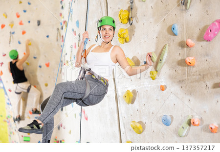 Sporty young girl in safety gear doing difficult wall climb in climbing gym Sporty young girl in safety gear doing difficult wall climb in climbing gym 137357217