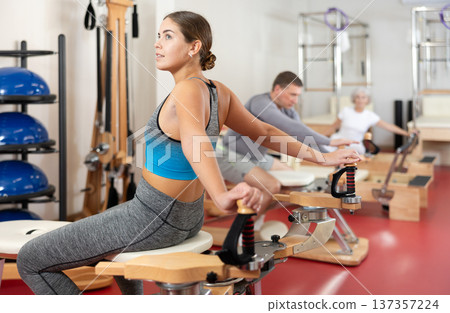 Young woman stretches her arms using pilates machine while sitting backwards 137357224