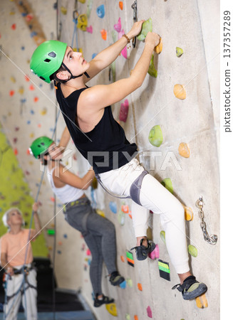 Well-equipped, positive young man training on stone wall, artificial mountain with rocks, climbing up in bouldering park indoors 137357289