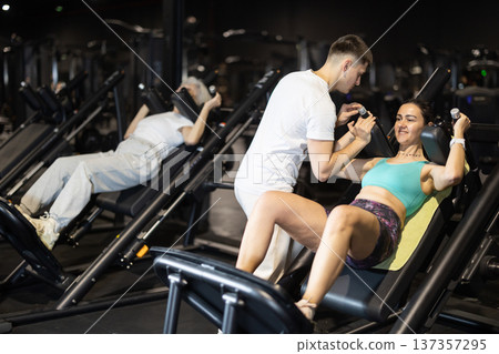 Woman doing hack squats with trainer support in gym Woman doing hack squats with trainer support in gym 137357295