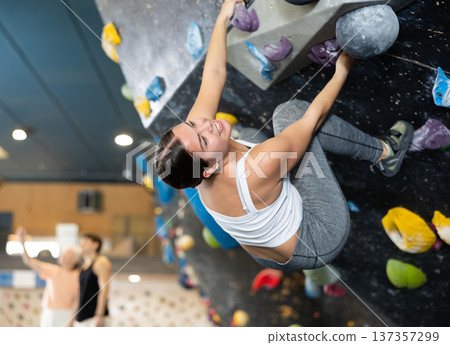 Female fitness climber training at bouldering gym. Young woman rise up on artificial climbing wall Female fitness climber training at bouldering gym. Young woman rise up on artificial climbing wall 137357299