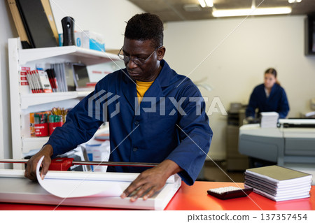Focused African American man in blue robe uniform trimming the printed work on the cutter in the typography 137357454