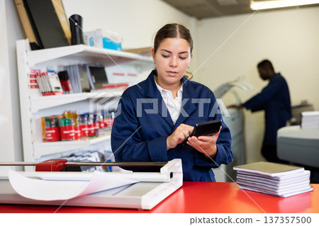 Focused young female typographer in uniform calculates the cost of printed notepads and sheet in the typography 137357500