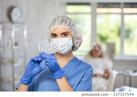 Nurse in blue uniform with mask and gloves behind hat holding syringe with medicine, patient behind. 137357544