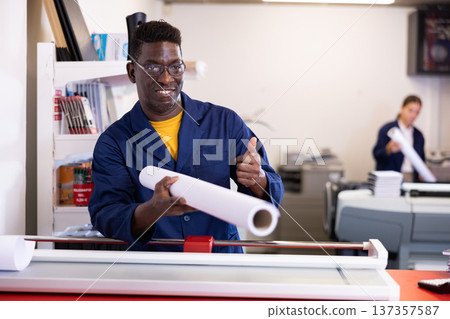 Excited middle-aged African American male typographer in uniform showing large format paper in a roll in the typography Excited middle-aged African American male typographer in uniform showing large format paper in a roll in the typography 137357587