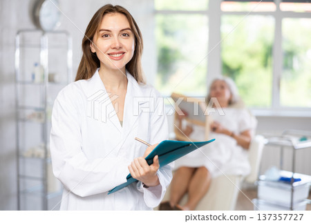 Woman surgeon in uniform is smiling and writing on clipboard in clinic with patient who lying behind 137357727