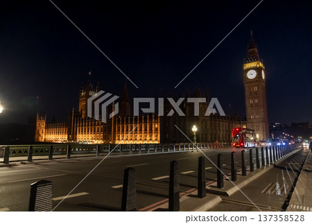 Elizabeth Tower and the Palace of Westminster illuminated at night beside a city road. A red double decker bus travels along the bridge under street lighting. 137358528