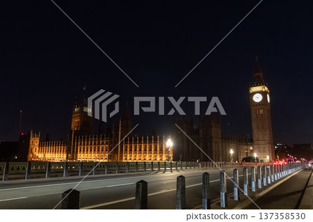 Elizabeth Tower and the Palace of Westminster illuminated at night beside a city road. A red double decker bus travels along the bridge under street lighting. 137358530