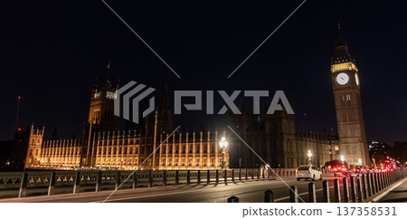 Elizabeth Tower and the Palace of Westminster illuminated at night beside a city road. A red double decker bus travels along the bridge under street lighting. 137358531