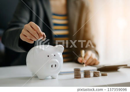 Business woman hand putting money coin into piggy bank with step of growing stack coins for saving money at modern office Business woman hand putting money coin into piggy bank with step of growing stack coins for saving money at modern office 137359096