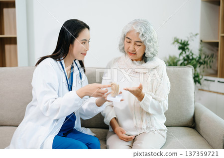 Female doctor explaining ear anatomy to elderly woman during home medical consultation. Hearing care, senior health, audiology, prevention, wellness and healthcare Female doctor explaining ear anatomy to elderly woman during home medical consultation. Hearing care, senior health, audiology, prevention, wellness and healthcare 137359721