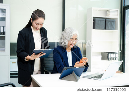 Senior and junior Asian businesswomen discussing documents in modern office, mentorship, teamwork, leadership 137359903
