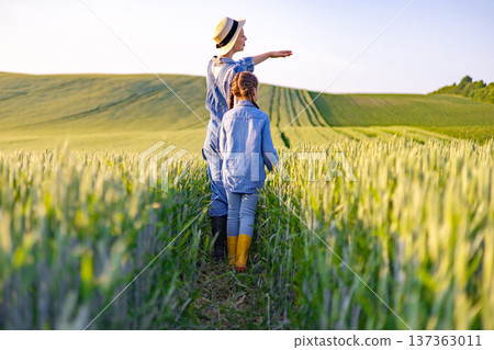 A woman in a straw hat and overalls points towards the horizon while walking with a young girl through a lush green wheat field at sunset A woman in a straw hat and overalls points towards the horizon while walking with a young girl through a lush green wheat field at sunset 137363011