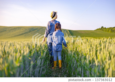 A mother and her daughter walk through a golden wheat field under a clear blue sky, enjoying a serene rural landscape 137363012
