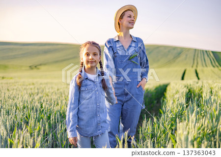 A woman in overalls and a straw hat stands with a young girl in a wheat field at sunset, both wearing denim shirts 137363043