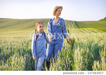 A mother and her young daughter walk hand-in-hand through a lush green wheat field under a clear sky, embodying a sense of rural life and family connection A mother and her young daughter walk hand-in-hand through a lush green wheat field under a clear sky, embodying a sense of rural life and family connection 137363057