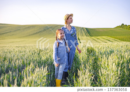 A woman and a young girl stand in a vast field of wheat, looking towards the horizon under a clear sky A woman and a young girl stand in a vast field of wheat, looking towards the horizon under a clear sky 137363058