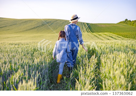 A farmer wearing overalls and a hat walks with a young child through a lush green wheat field under a clear sky 137363062