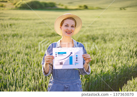 A smiling woman in a straw hat and overalls holds a paper with a growing bar chart and arrow in a field of green crops A smiling woman in a straw hat and overalls holds a paper with a growing bar chart and arrow in a field of green crops 137363070