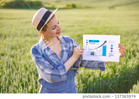 A woman in a straw hat and overalls stands in a field of green crops, holding up a paper with a rising bar graph and statistics 137363074