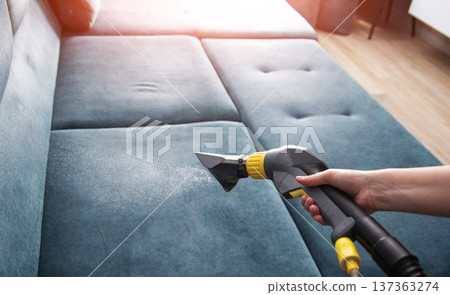 One person hand holding cleaning tool spraying water on blue fabric sofa. Professional wet cleaning service of upholstery furniture using extractor machine at home. One person hand holding cleaning tool spraying water on blue fabric sofa. Professional wet cleaning service of upholstery furniture using extractor machine at home. 137363274