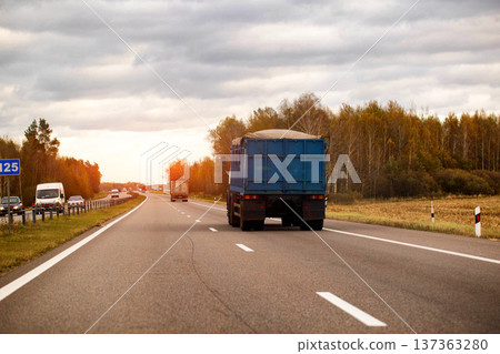 Blue truck driving on highway at sunset, autumn forest and field beside road, vehicles on multi-lane pavement, transportation of goods on open road under cloudy sky 137363280