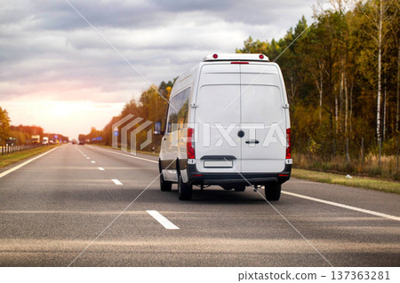 White cargo van driving on highway at sunset, autumn trees line road, delivery vehicle traveling on asphalt route during golden hour with cloudy sky 137363281