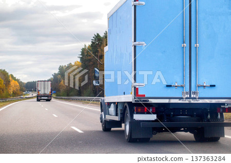Blue delivery truck rear view driving on multi-lane highway during day with autumn color trees lining roadside under overcast sky freight transportation logistics commerce distribution service 137363284