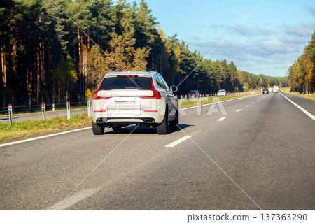 Silver SUV traveling on highway beside forest in autumn day, vehicle movement on asphalt road with trees and blue sky, transportation on open road Silver SUV traveling on highway beside forest in autumn day, vehicle movement on asphalt road with trees and blue sky, transportation on open road 137363290